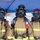 USS Hampton Conducts Fire Prevention Drill Three firefighters in full personal protective equipment stand together under a blue canopy tent, facing the camera. All three are wearing tan/gold turnout gear with yellow reflective striping, full-face respirator masks, and helmets. Their gear is marked with handwritten identifiers and the number '767', and they are equipped with SCBA (Self-Contained Breathing Apparatus) units, radios, and flashlights.