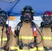 Three firefighters in full personal protective equipment stand together under a blue canopy tent, facing the camera. All three are wearing tan/gold turnout gear with yellow reflective striping, full-face respirator masks, and helmets. Their gear is marked with handwritten identifiers and the number '767', and they are equipped with SCBA (Self-Contained Breathing Apparatus) units, radios, and flashlights.