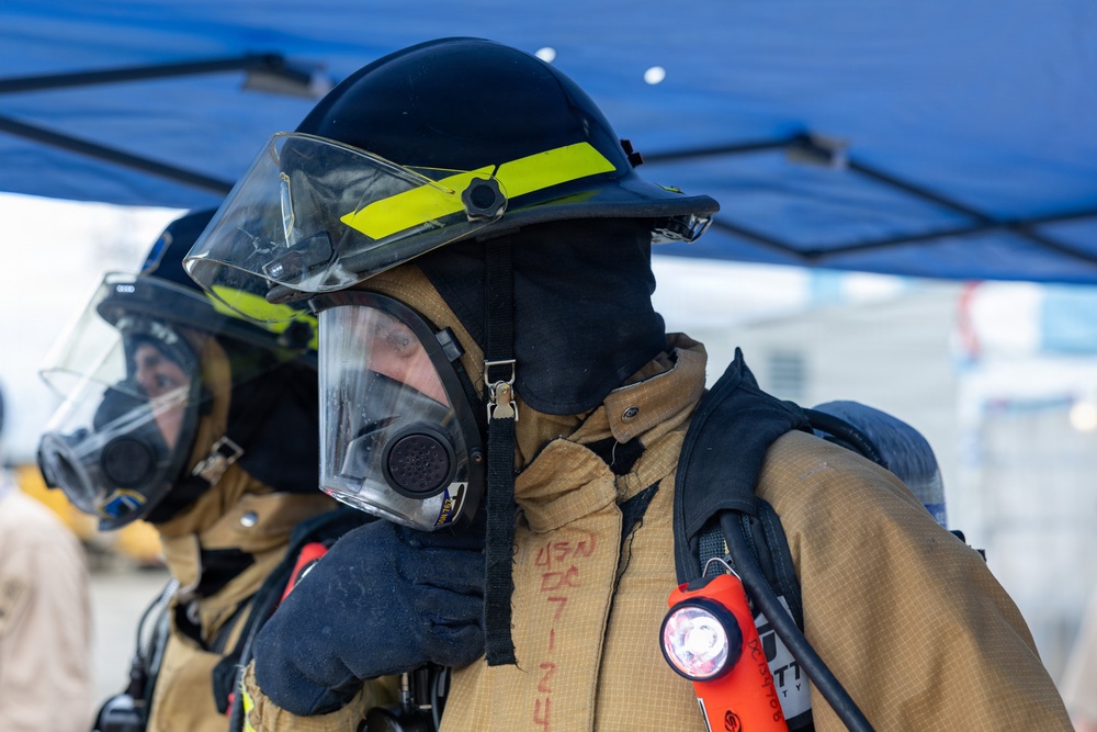 Two firefighters are shown in full personal protective equipment (PPE), including turnout gear, SCBA face masks, and helmets with yellow reflective striping. The foreground firefighter is adjusting or checking their SCBA mask while wearing a black hood and tan bunker coat. An orange Streamlight flashlight is clipped to the foreground firefighter's gear, and a blue canopy tent is visible in the background.