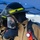USS Hampton Conducts Fire Prevention Drill Two firefighters are shown in full personal protective equipment (PPE), including turnout gear, SCBA face masks, and helmets with yellow reflective striping. The foreground firefighter is adjusting or checking their SCBA mask while wearing a black hood and tan bunker coat. An orange Streamlight flashlight is clipped to the foreground firefighter's gear, and a blue canopy tent is visible in the background.
