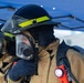 Two firefighters are shown in full personal protective equipment (PPE), including turnout gear, SCBA face masks, and helmets with yellow reflective striping. The foreground firefighter is adjusting or checking their SCBA mask while wearing a black hood and tan bunker coat. An orange Streamlight flashlight is clipped to the foreground firefighter's gear, and a blue canopy tent is visible in the background.