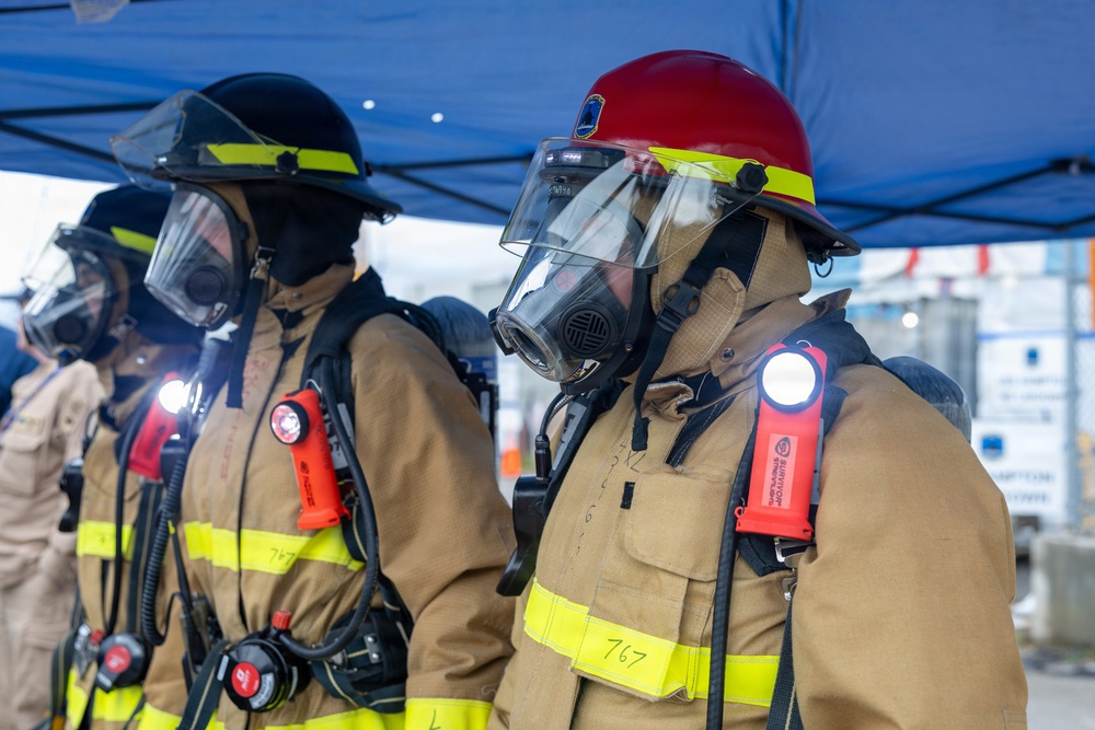The image shows multiple firefighters in full personal protective equipment (PPE) standing in formation under a blue canopy tent. They are wearing turnout gear, full-face respirator masks, SCBA (Self-Contained Breathing Apparatus) equipment, and helmets in black and red. The setting appears to be an outdoor staging area, possibly during a drill, training exercise, or pre-deployment briefing.