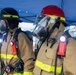 The image shows multiple firefighters in full personal protective equipment (PPE) standing in formation under a blue canopy tent. They are wearing turnout gear, full-face respirator masks, SCBA (Self-Contained Breathing Apparatus) equipment, and helmets in black and red. The setting appears to be an outdoor staging area, possibly during a drill, training exercise, or pre-deployment briefing.