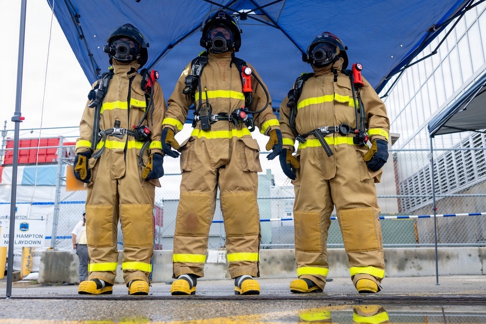 Three firefighters in full personal protective equipment stand side by side under a blue canopy tent, facing the camera in a formal pose. They are wearing tan/gold turnout gear with yellow reflective striping, full-face respirator masks, black helmets, SCBA (self-contained breathing apparatus) tanks on their backs, and yellow rubber boots. A sign partially visible in the background references 'USS Hampton' and 'Mold Laydown,' suggesting a naval or shipyard setting.