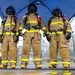 Three firefighters in full personal protective equipment stand side by side under a blue canopy tent, facing the camera in a formal pose. They are wearing tan/gold turnout gear with yellow reflective striping, full-face respirator masks, black helmets, SCBA (self-contained breathing apparatus) tanks on their backs, and yellow rubber boots. A sign partially visible in the background references 'USS Hampton' and 'Mold Laydown,' suggesting a naval or shipyard setting.