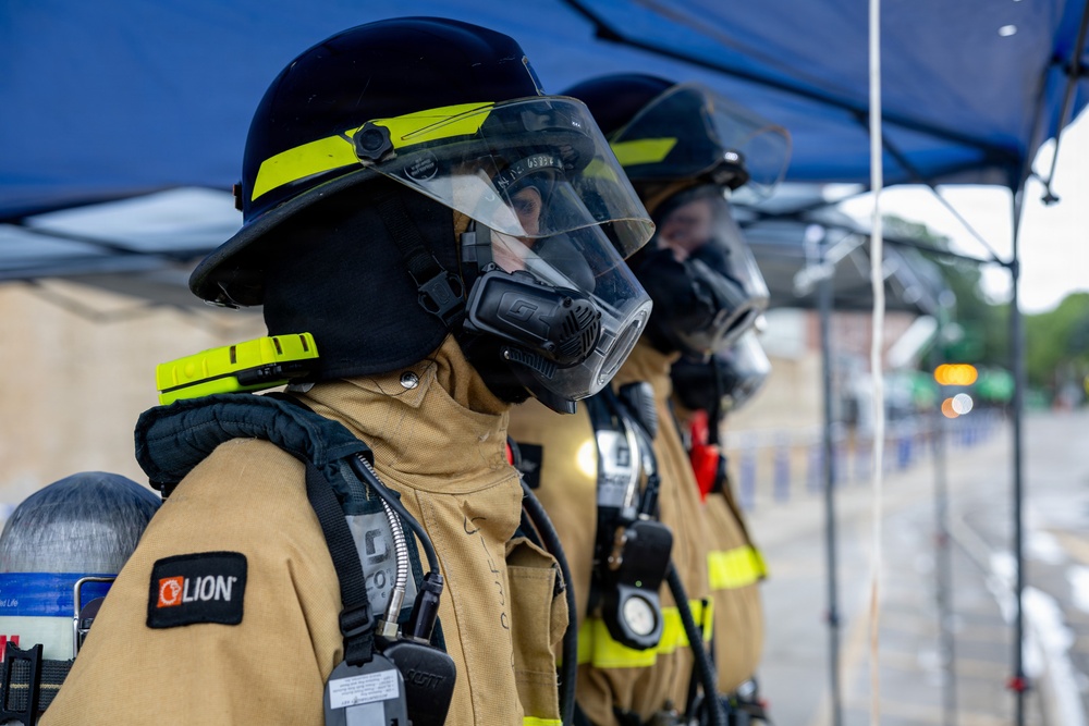Three firefighters in full personal protective equipment stand in a line under a blue canopy tent. They are wearing LION brand turnout gear, full-face respirator masks with face shields, black helmets with yellow-green reflective trim, and SCBA (Self-Contained Breathing Apparatus) tanks on their backs. The image appears to capture a training exercise, drill, or pre-deployment staging scenario.