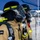 USS Hampton Conducts Fire Prevention Drill Three firefighters in full personal protective equipment stand in a line under a blue canopy tent. They are wearing LION brand turnout gear, full-face respirator masks with face shields, black helmets with yellow-green reflective trim, and SCBA (Self-Contained Breathing Apparatus) tanks on their backs. The image appears to capture a training exercise, drill, or pre-deployment staging scenario.