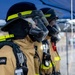 Three firefighters in full personal protective equipment stand in a line under a blue canopy tent. They are wearing LION brand turnout gear, full-face respirator masks with face shields, black helmets with yellow-green reflective trim, and SCBA (Self-Contained Breathing Apparatus) tanks on their backs. The image appears to capture a training exercise, drill, or pre-deployment staging scenario.