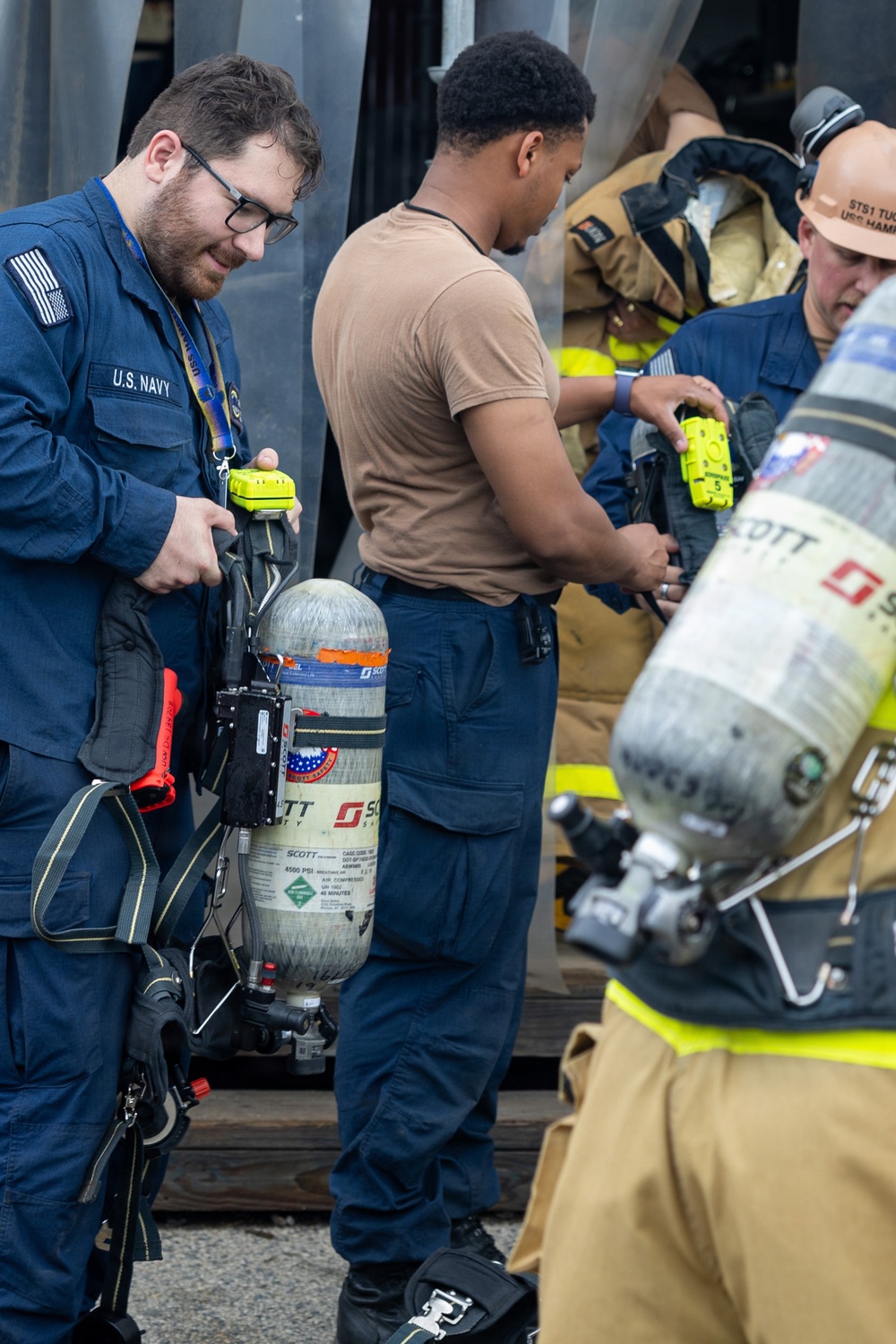 U.S. Navy personnel are preparing or inspecting Self-Contained Breathing Apparatus (SCBA) equipment, likely during a damage control or firefighting drill. At least one sailor is wearing a U.S. Navy uniform with a name tag, while others are in various stages of donning firefighting gear. The scene appears to be on or near a naval vessel based on the metal structural elements visible in the background.