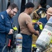 U.S. Navy personnel are preparing or inspecting Self-Contained Breathing Apparatus (SCBA) equipment, likely during a damage control or firefighting drill. At least one sailor is wearing a U.S. Navy uniform with a name tag, while others are in various stages of donning firefighting gear. The scene appears to be on or near a naval vessel based on the metal structural elements visible in the background.