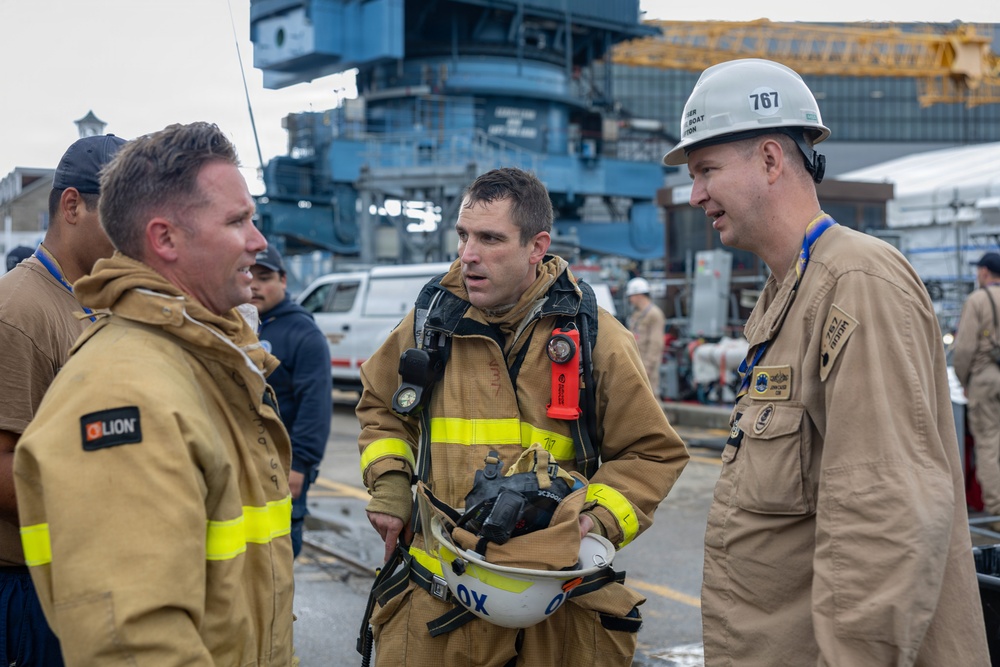Three individuals in protective work gear are engaged in conversation at what appears to be a naval shipyard or industrial port facility. The central figure is wearing full firefighting turnout gear with SCBA equipment, while the others wear tan coveralls. A large naval vessel or industrial structure is visible in the background.