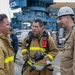 Three individuals in protective work gear are engaged in conversation at what appears to be a naval shipyard or industrial port facility. The central figure is wearing full firefighting turnout gear with SCBA equipment, while the others wear tan coveralls. A large naval vessel or industrial structure is visible in the background.