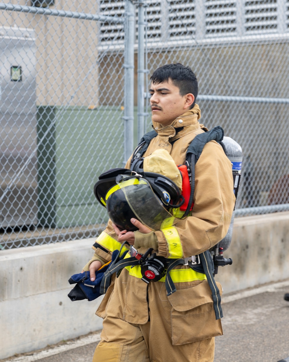 A young firefighter in full turnout gear is carrying his helmet and SCBA (Self-Contained Breathing Apparatus) equipment while walking near a chain-link fence and concrete barrier. He is wearing a tan/gold structural firefighting suit with yellow reflective trim and has an air cylinder mounted on his back. The setting appears to be a training facility or fire station exterior.