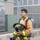 USS Hampton Conducts Fire Prevention Drill A young firefighter in full turnout gear is carrying his helmet and SCBA (Self-Contained Breathing Apparatus) equipment while walking near a chain-link fence and concrete barrier. He is wearing a tan/gold structural firefighting suit with yellow reflective trim and has an air cylinder mounted on his back. The setting appears to be a training facility or fire station exterior.