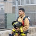 A young firefighter in full turnout gear is carrying his helmet and SCBA (Self-Contained Breathing Apparatus) equipment while walking near a chain-link fence and concrete barrier. He is wearing a tan/gold structural firefighting suit with yellow reflective trim and has an air cylinder mounted on his back. The setting appears to be a training facility or fire station exterior.