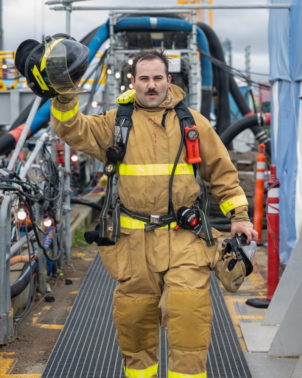 A firefighter in full turnout gear walks forward on a metal grating walkway in an industrial setting. He is holding his helmet raised in his left hand and carrying a face mask/respirator in his right hand. The gear includes SCBA harness, high-visibility yellow reflective strips, and an orange flashlight clipped to his chest.