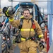 A firefighter in full turnout gear walks forward on a metal grating walkway in an industrial setting. He is holding his helmet raised in his left hand and carrying a face mask/respirator in his right hand. The gear includes SCBA harness, high-visibility yellow reflective strips, and an orange flashlight clipped to his chest.