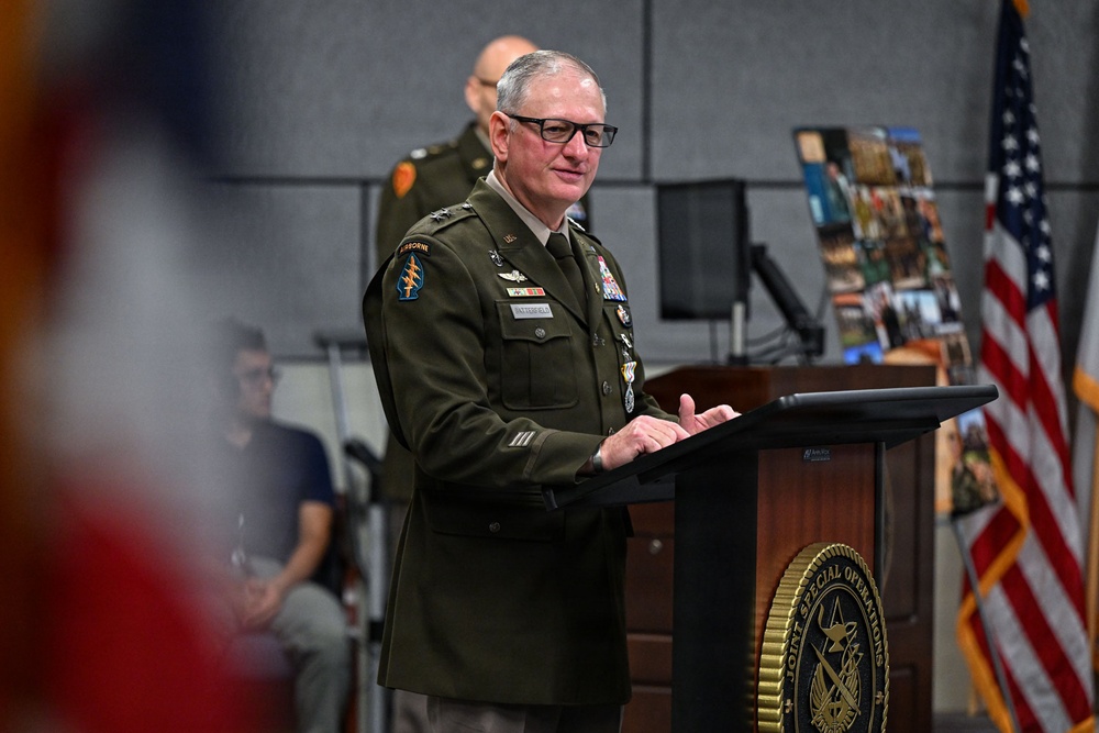 A senior U.S. Army officer in dress green uniform is speaking at a podium bearing the seal of Joint Special Operations Command (JSOC). The officer displays multiple medals, badges, and unit patches including an Airborne tab and what appears to be a Special Forces patch. An American flag is visible to the right, and a second uniformed soldier stands in the background.