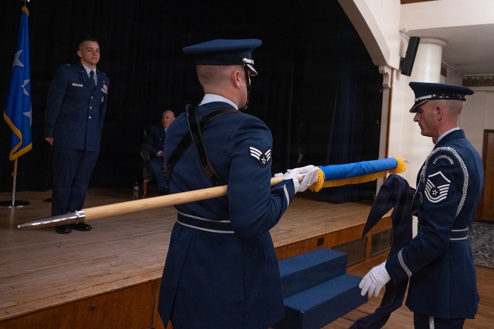 A formal U.S. Air Force military ceremony is taking place in what appears to be an auditorium or ceremonial hall. Honor Guard members in dress blue uniforms are handling a guidon or unit flag on a staff, while an officer stands at attention on a raised stage. The scene is consistent with a change of command, activation, or inactivation ceremony.