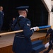A formal U.S. Air Force military ceremony is taking place in what appears to be an auditorium or ceremonial hall. Honor Guard members in dress blue uniforms are handling a guidon or unit flag on a staff, while an officer stands at attention on a raised stage. The scene is consistent with a change of command, activation, or inactivation ceremony.