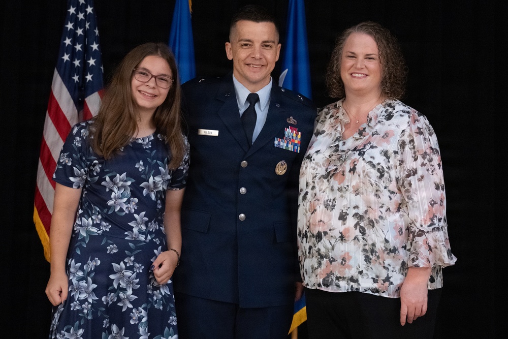 A U.S. Air Force officer in dress blues uniform poses with two civilians, likely family members, in front of an American flag and Air Force flag. The officer's name tag reads 'WILSON' and he wears multiple service ribbons and insignia. This appears to be a formal military ceremony or promotion event.