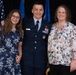 A U.S. Air Force officer in dress blues uniform poses with two civilians, likely family members, in front of an American flag and Air Force flag. The officer's name tag reads 'WILSON' and he wears multiple service ribbons and insignia. This appears to be a formal military ceremony or promotion event.
