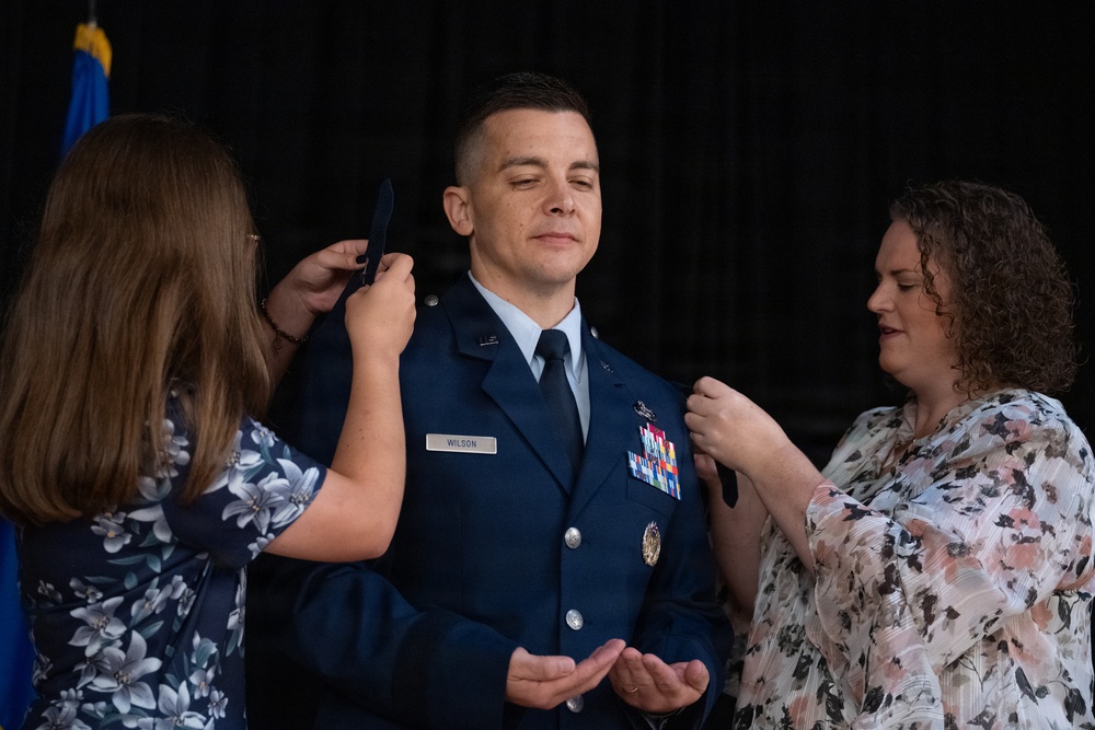 A U.S. Air Force officer named Wilson is undergoing a promotion ceremony, with two women pinning new rank insignia onto the shoulders of his dress blue uniform. The officer stands with eyes closed and hands open in a ceremonial posture while the women work on both shoulders simultaneously. A blue military flag is partially visible in the background.