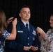 A U.S. Air Force officer named Wilson is undergoing a promotion ceremony, with two women pinning new rank insignia onto the shoulders of his dress blue uniform. The officer stands with eyes closed and hands open in a ceremonial posture while the women work on both shoulders simultaneously. A blue military flag is partially visible in the background.