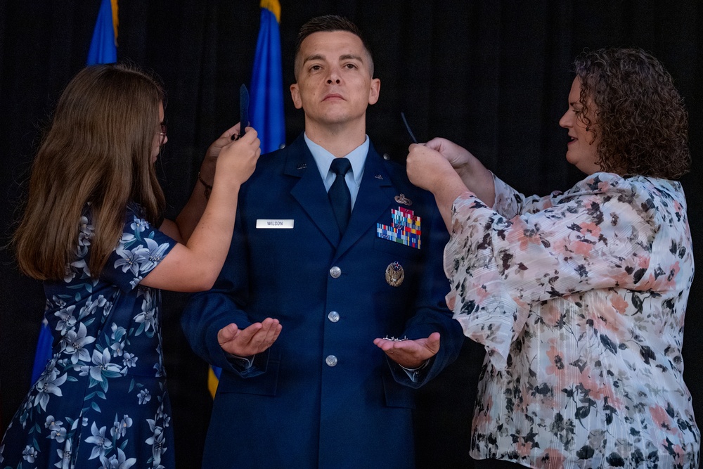 A U.S. Air Force officer named Wilson is undergoing a promotion ceremony, with two women pinning new rank insignia onto the epaulettes of his dress blue uniform. The officer stands at attention with palms open while family members or colleagues attach the new rank devices to both shoulders simultaneously. A blue U.S. Air Force flag is visible in the background.