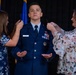 A U.S. Air Force officer named Wilson is undergoing a promotion ceremony, with two women pinning new rank insignia onto the epaulettes of his dress blue uniform. The officer stands at attention with palms open while family members or colleagues attach the new rank devices to both shoulders simultaneously. A blue U.S. Air Force flag is visible in the background.