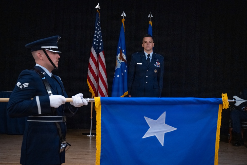A formal U.S. Air Force military ceremony is taking place, likely a promotion or change of command event. A one-star general officer flag (Brigadier General) is being displayed in the foreground by an Airman in dress blues with white gloves. An officer stands at attention in the background flanked by the U.S. flag and Air Force flag.