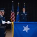 A formal U.S. Air Force military ceremony is taking place, likely a promotion or change of command event. A one-star general officer flag (Brigadier General) is being displayed in the foreground by an Airman in dress blues with white gloves. An officer stands at attention in the background flanked by the U.S. flag and Air Force flag.