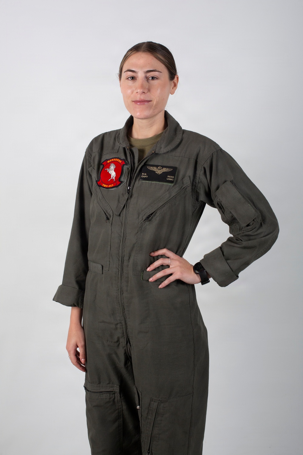 A person wearing an olive green military flight suit stands against a neutral gray background in a portrait-style photograph. The flight suit displays a squadron patch on the left chest reading 'WARHORSE HMH-465' and a name tag on the right chest reading 'R.N. CAPT REED USMC' with aviator wings above it. The subject has hair pulled back and stands with one hand on their hip.