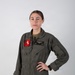 A person wearing an olive green military flight suit stands against a neutral gray background in a portrait-style photograph. The flight suit displays a squadron patch on the left chest reading 'WARHORSE HMH-465' and a name tag on the right chest reading 'R.N. CAPT REED USMC' with aviator wings above it. The subject has hair pulled back and stands with one hand on their hip.