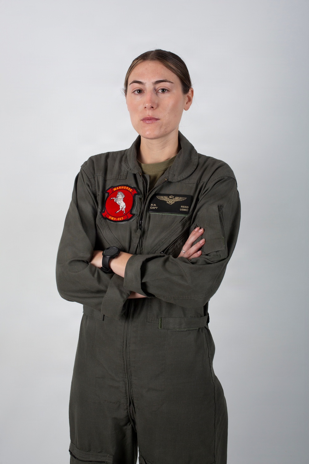 A formal portrait of a U.S. Marine Corps officer in an olive drab flight suit, standing with arms crossed against a neutral gray background. The subject wears squadron patch 'HMH-465 Warhorse' on the left chest and a naval aviator wings badge on the right chest. Name tape reads 'REED' with rank 'CAPT' and branch 'USMC' visible.