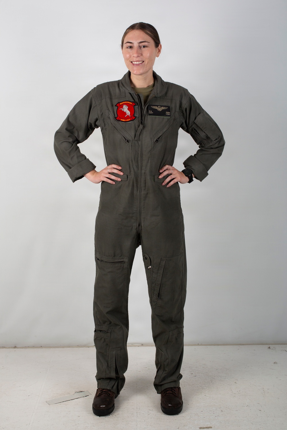 A female military aviator stands in a confident pose with hands on hips against a neutral white/grey background. She is wearing an olive green flight suit with visible unit patch and name tag, consistent with U.S. Marine Corps aviation attire. The image appears to be an official or semi-official portrait photograph.