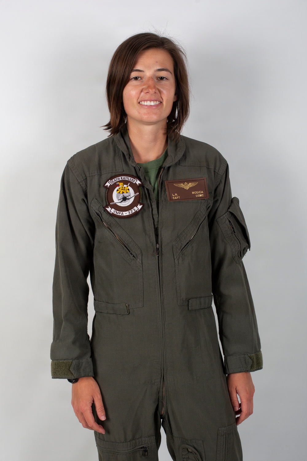 A person wearing an olive green military flight suit stands against a neutral gray background in what appears to be an official portrait photograph. The flight suit displays a squadron patch reading 'Death Rattlers VMFA-323' and a name tag reading 'L.N. Modica, CAPT, USMC' with naval aviator wings. The subject is smiling and facing the camera directly.
