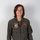 A person wearing an olive green military flight suit stands against a neutral gray background in what appears to be an official portrait photograph. The flight suit displays a squadron patch reading 'Death Rattlers VMFA-323' and a name tag reading 'L.N. Modica, CAPT, USMC' with naval aviator wings. The subject is smiling and facing the camera directly.