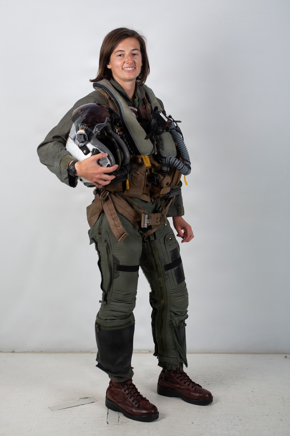 A person is photographed in a full military flight suit against a neutral light gray background in what appears to be an official portrait setting. They are wearing a complete aviator ensemble including a green flight suit, tactical vest with harness, G-suit pants, and brown flight boots. They are holding a flight helmet and smiling at the camera.
