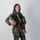 A person is photographed in a full military flight suit against a neutral light gray background in what appears to be an official portrait setting. They are wearing a complete aviator ensemble including a green flight suit, tactical vest with harness, G-suit pants, and brown flight boots. They are holding a flight helmet and smiling at the camera.