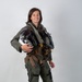 A person is photographed in a full military flight suit against a neutral light gray background in what appears to be an official portrait setting. They are wearing a complete aviator ensemble including a green flight suit, tactical vest with harness, G-suit pants, and brown flight boots. They are holding a flight helmet and smiling at the camera.