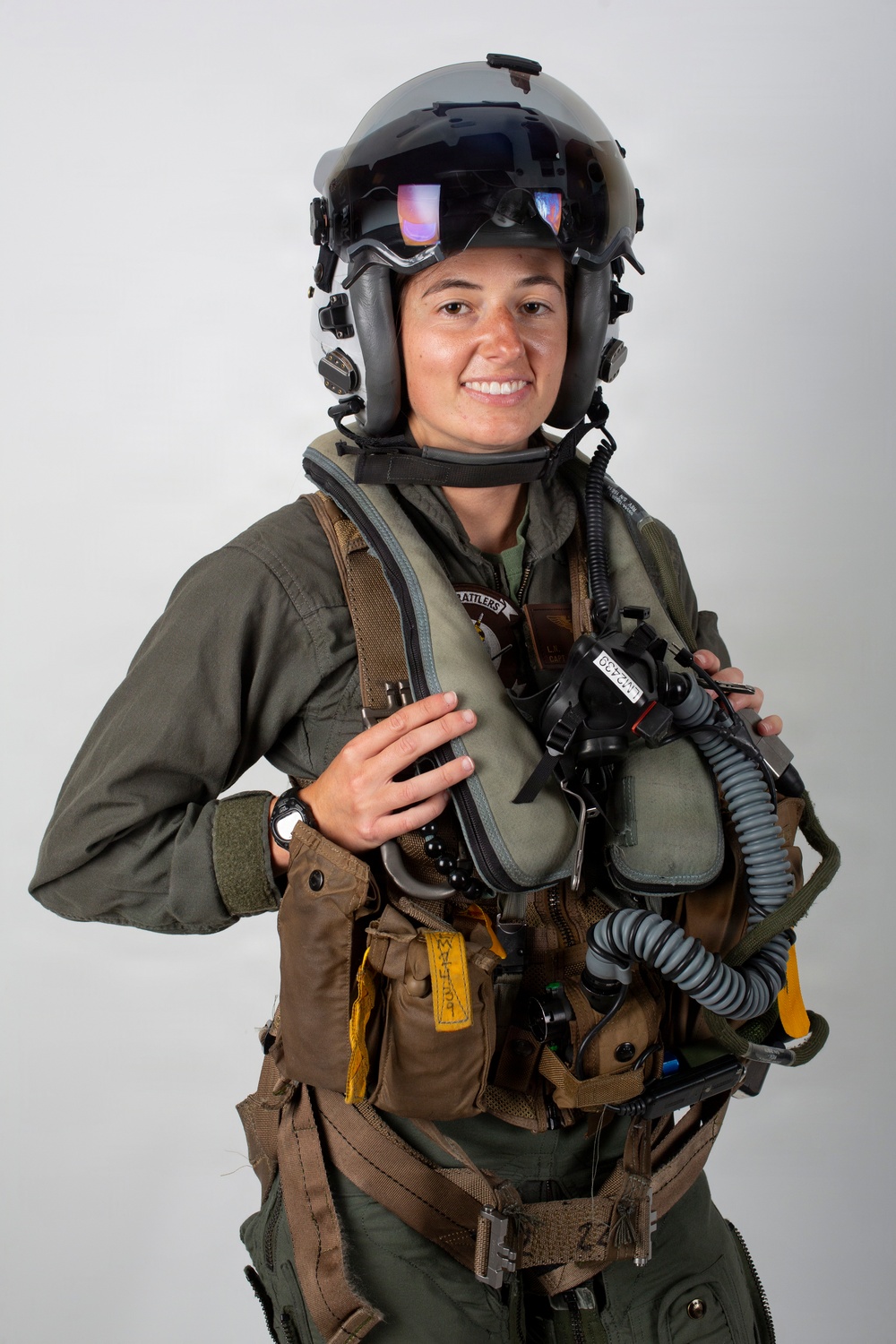 A military aviator is photographed against a plain light gray background in a formal portrait-style image. The individual is wearing full flight gear including a helmet with visor, flight suit, survival vest, and oxygen mask assembly. The subject is smiling and appears to be posing for an official or promotional photograph.