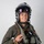 A military aviator is photographed against a plain light gray background in a formal portrait-style image. The individual is wearing full flight gear including a helmet with visor, flight suit, survival vest, and oxygen mask assembly. The subject is smiling and appears to be posing for an official or promotional photograph.