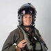 A military aviator is photographed against a plain light gray background in a formal portrait-style image. The individual is wearing full flight gear including a helmet with visor, flight suit, survival vest, and oxygen mask assembly. The subject is smiling and appears to be posing for an official or promotional photograph.