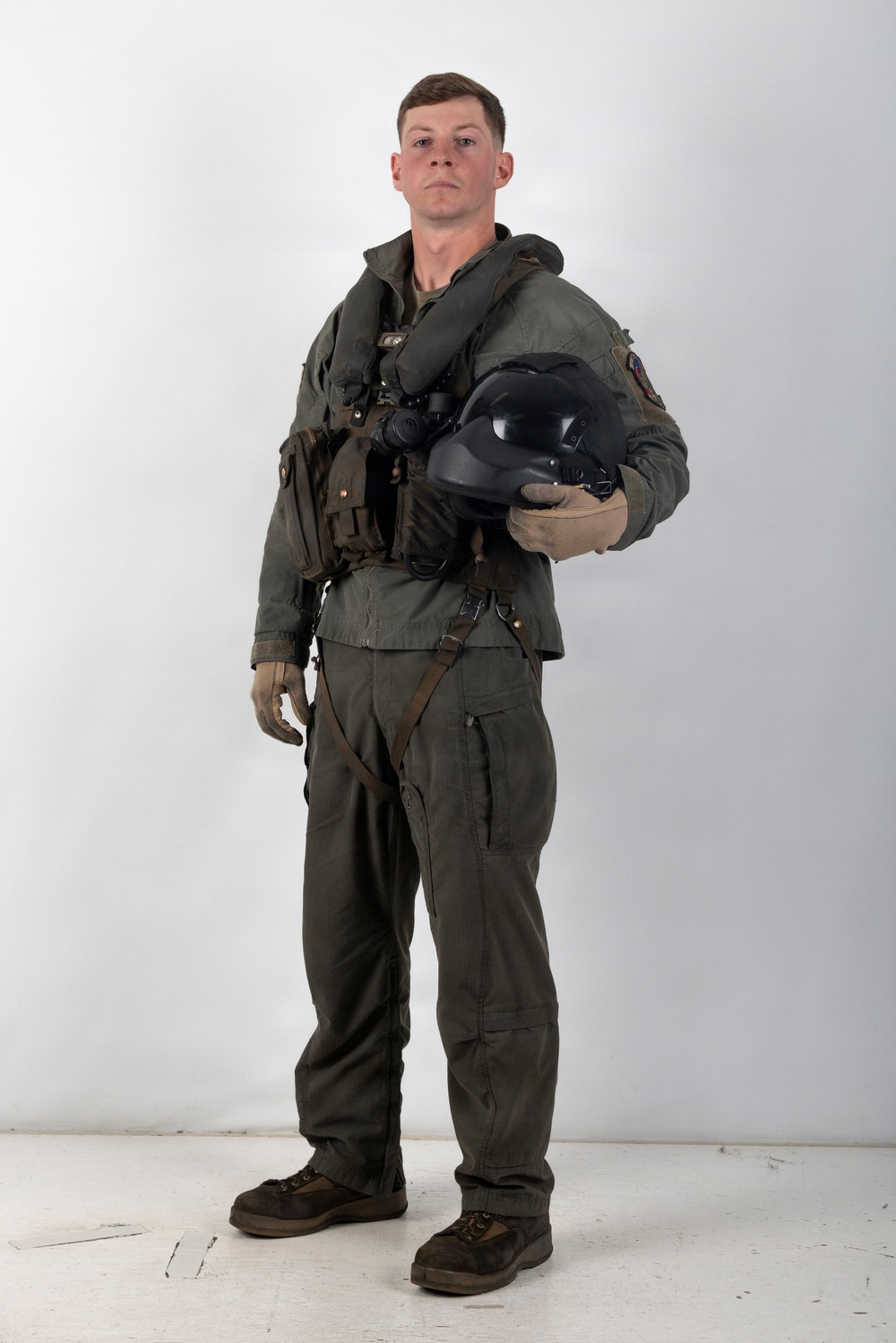 A male individual stands in a studio setting against a white/light gray background, wearing a full military flight suit ensemble. He is holding a black flight helmet under his right arm and is equipped with a survival vest, harness straps, and tactical gear. The image appears to be a formal documentation or reference photograph of military aviation equipment.
