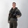MCAS Marines pose for content 2026 A male individual stands in a studio setting against a white/light gray background, wearing a full military flight suit ensemble. He is holding a black flight helmet under his right arm and is equipped with a survival vest, harness straps, and tactical gear. The image appears to be a formal documentation or reference photograph of military aviation equipment.