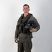 A male individual stands in a studio setting against a white/light gray background, wearing a full military flight suit ensemble. He is holding a black flight helmet under his right arm and is equipped with a survival vest, harness straps, and tactical gear. The image appears to be a formal documentation or reference photograph of military aviation equipment.