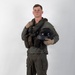 A young male military aviator stands in a full-body portrait pose against a plain white/light gray studio backdrop. He is wearing a complete flight suit ensemble with survival/tactical vest, harness, and is holding a flight helmet. The image appears to be a professional or official military portrait photograph.