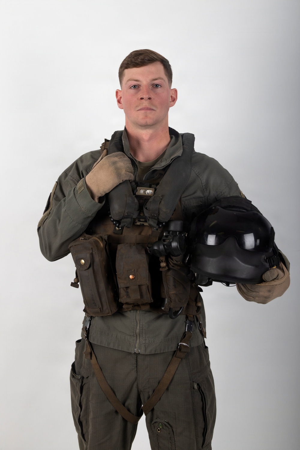 A young male military aviator is posed against a plain light gray studio background in a formal portrait-style photograph. He is wearing a full flight suit ensemble including a survival vest, harness, and gloves, and is holding a flight helmet in his right hand. The image appears to be an official or professional military portrait.