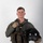 MCAS Marines pose for content 2026 A young male military aviator is posed against a plain light gray studio background in a formal portrait-style photograph. He is wearing a full flight suit ensemble including a survival vest, harness, and gloves, and is holding a flight helmet in his right hand. The image appears to be an official or professional military portrait.